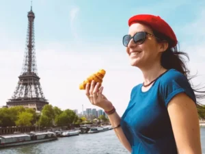 A woman in a red beret smiling while holding a croissant with the Eiffel Tower in the background, symbolizing French cultural immersion.
