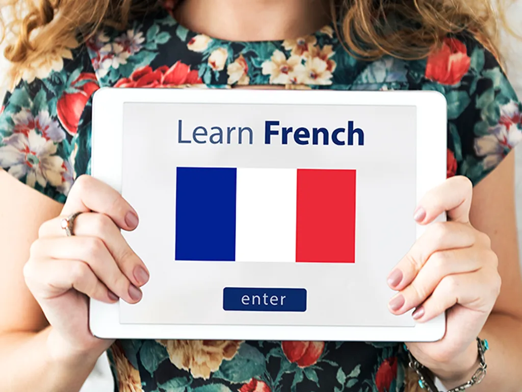 A person wearing a floral shirt holding a tablet that displays the text "Learn French," the French flag, and an "enter" button on its screen.