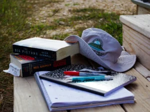 A collection of study materials, including books, notebooks, pens, and a bucket hat, resting on a wooden bench outdoors, ready for a study session.