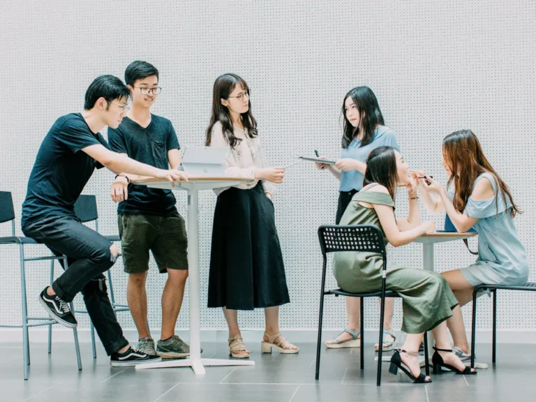 A group of six young students are collaborating around high-top tables, some standing and some sitting, engaged in a study session.